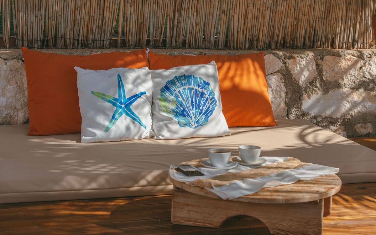 A cushioned outdoor daybed with colorful pillows and a small wooden table with two coffee cups in the sun.