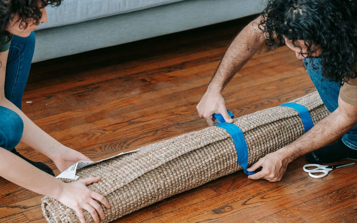 A couple wrapping a rolled carpet with adhesive tape, ready for moving.