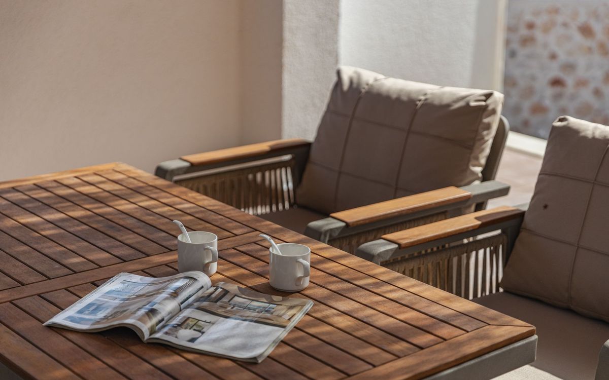 Two cushioned armchairs with wood armrests beside a slatted wooden table with a magazine and two coffee cups in morning sun.