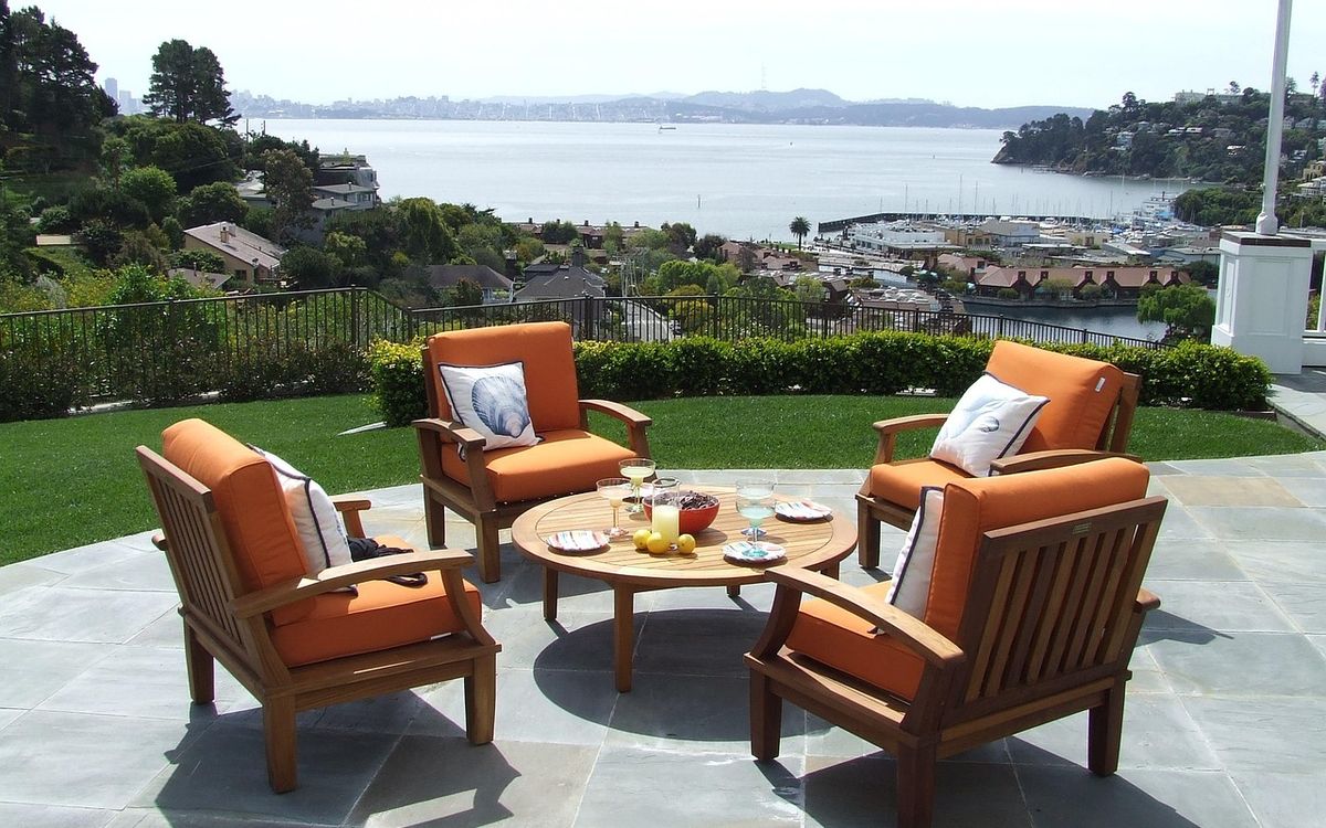 Teak patio armchairs with orange cushions around a round coffee table on a stone terrace overlooking the water.
