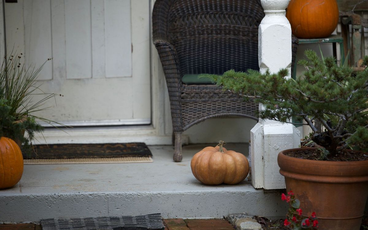 Dark wicker chair on a front porch beside pumpkins and a potted evergreen.