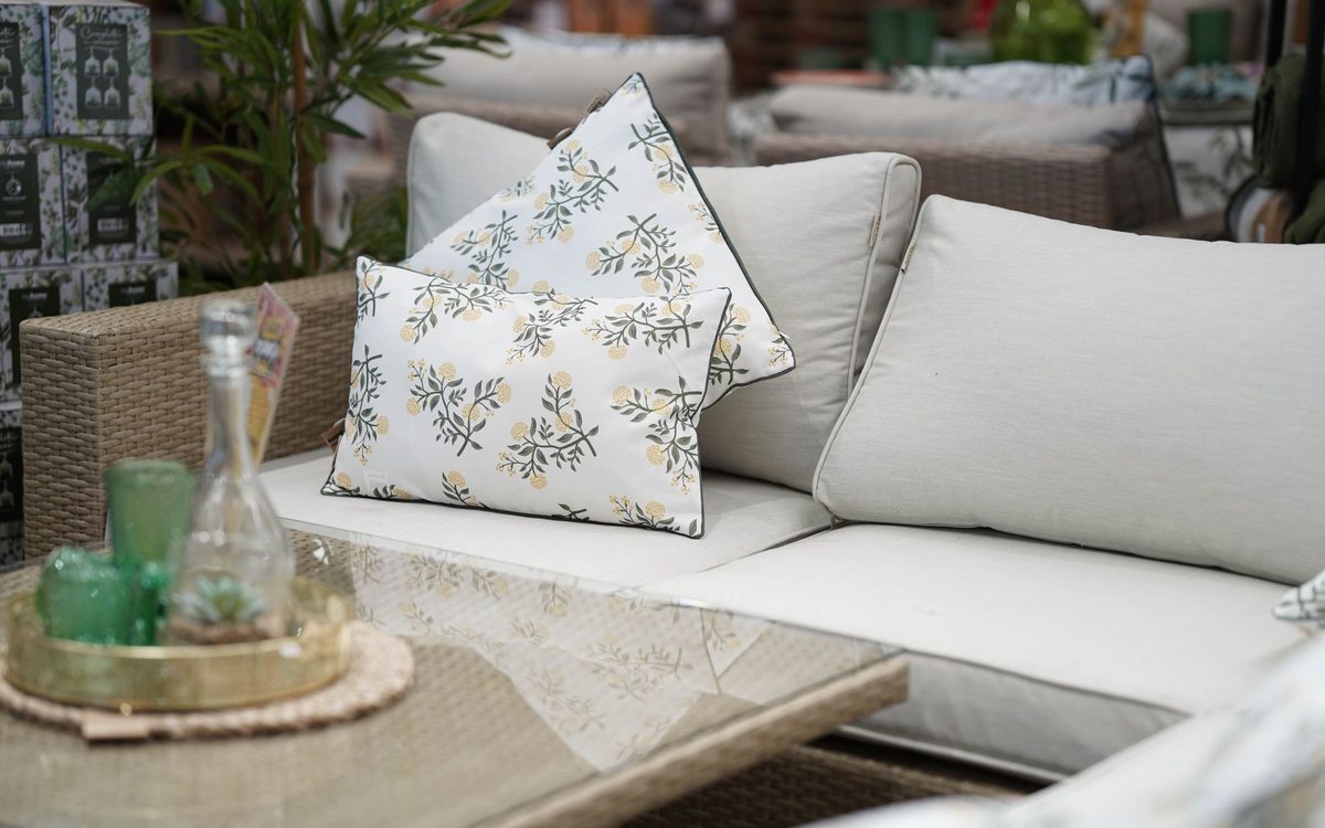 Wicker sofa with beige cushions and floral throw pillows behind a glass-top table holding a tray of green glassware.