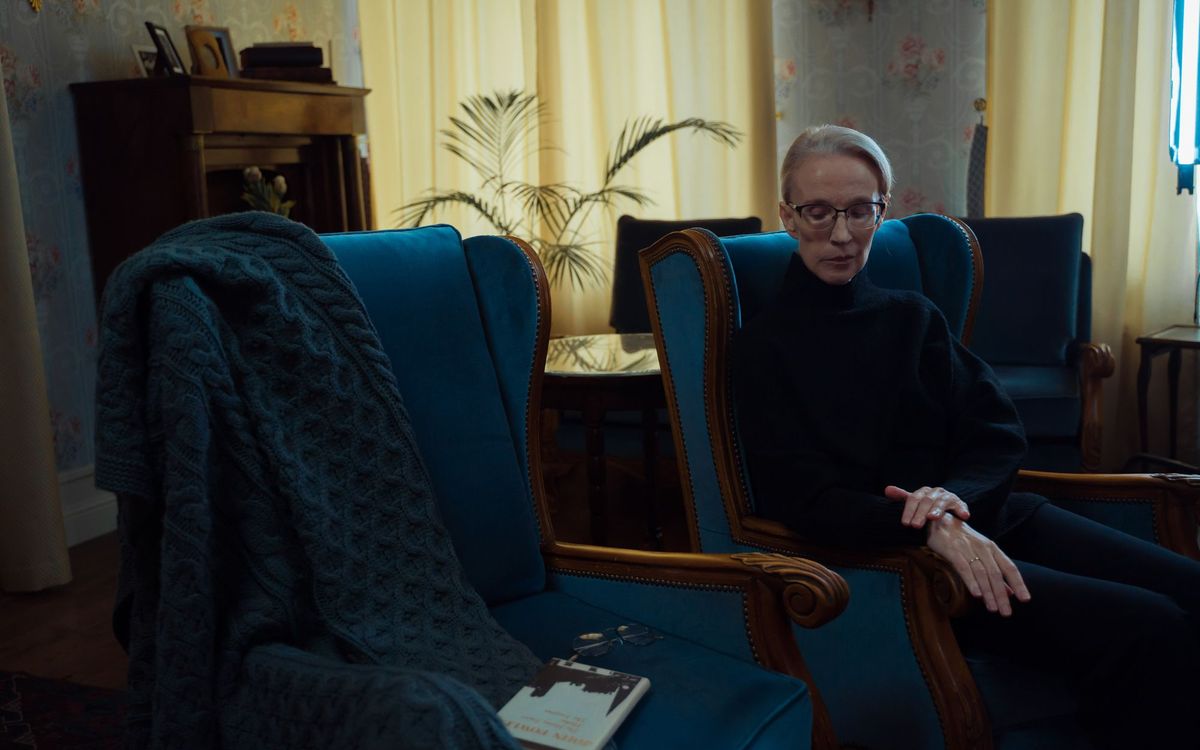 Elderly woman with glasses sitting in a blue wingback armchair in a classic living room.