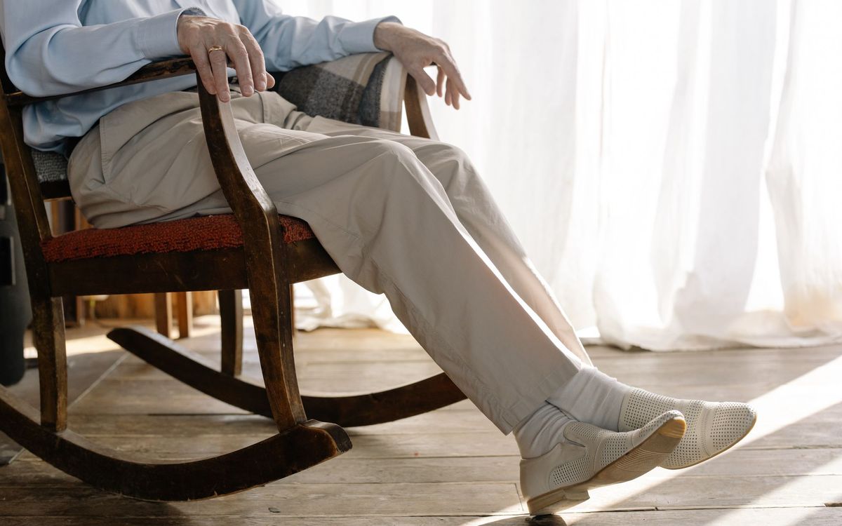Older man seated in a wooden rocking chair beside sheer curtains, feet resting on the floor.