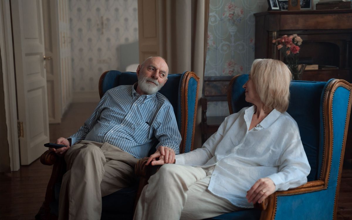 Older couple sitting side by side in blue armchairs, holding hands in a living room.