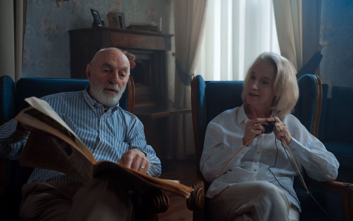 Older couple sitting in armchairs at home, one reading a newspaper and one knitting
