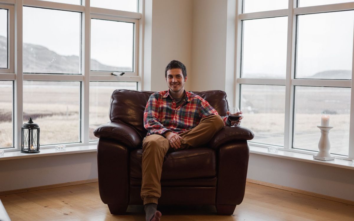 A man in a plaid shirt relaxing in a dark leather armchair in front of large windows, with open floor around the chair