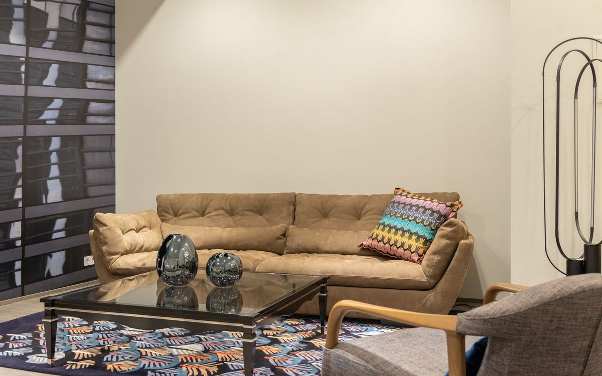 Living room with a tan sofa against a plain wall, a glass coffee table and a gray armchair in the foreground