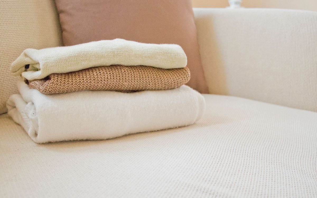 Stack of three folded knit sweaters and blankets in cream and tan resting on a cream fabric couch