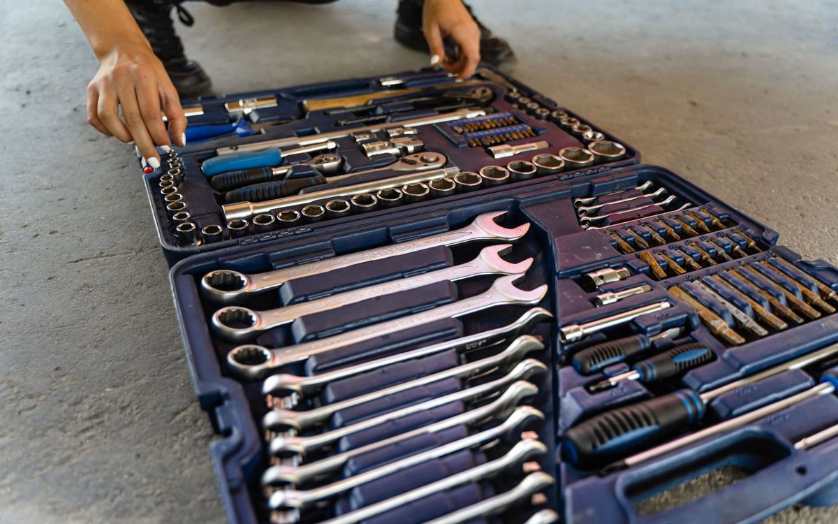Open blue toolbox on a concrete floor with wrenches, sockets and screwdrivers laid out, and a hand reaching in