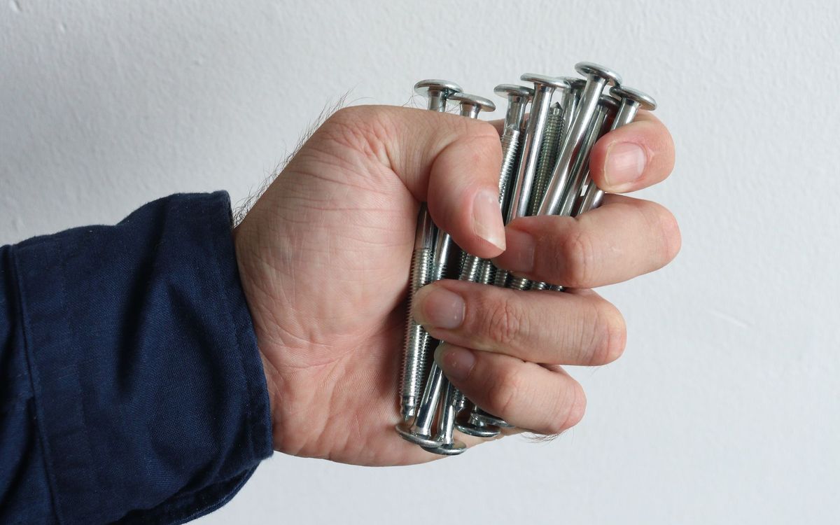Hand gripping a bunch of long shiny metal bolts against a plain white wall