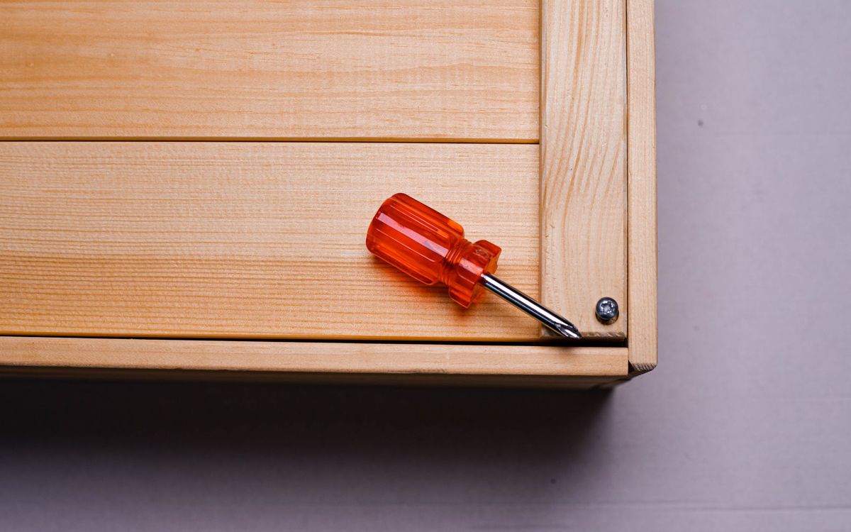 Red-handled screwdriver resting on the corner of a wooden box next to a screw