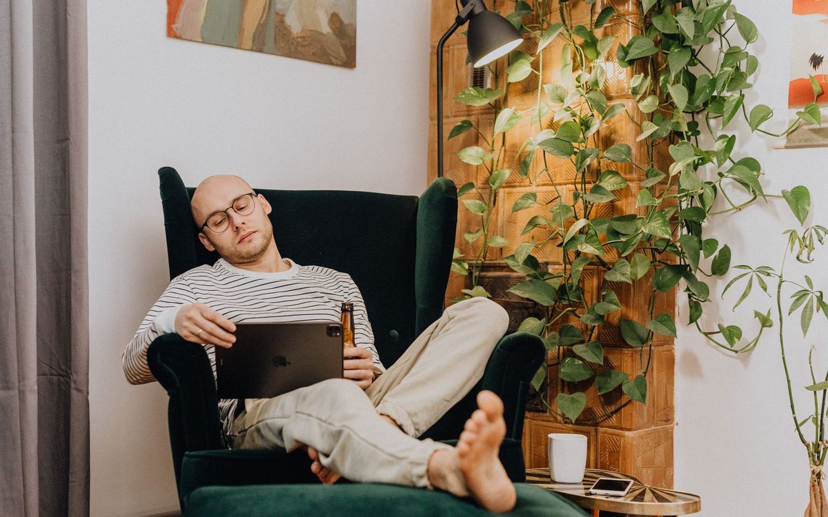 A man in a striped top reclines barefoot in a dark green armchair with a tablet and a footstool in a living room with plants