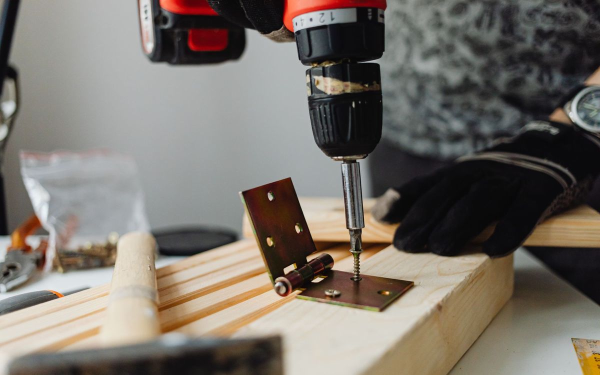 Gloved hand using a cordless drill to drive a screw into a metal hinge on a wooden block, showing the fastener work involved in taking furniture apart