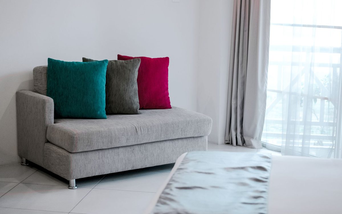 Small grey sofa with teal, grey and red cushions in a bright room next to a window, a stand-in for a recliner sofa