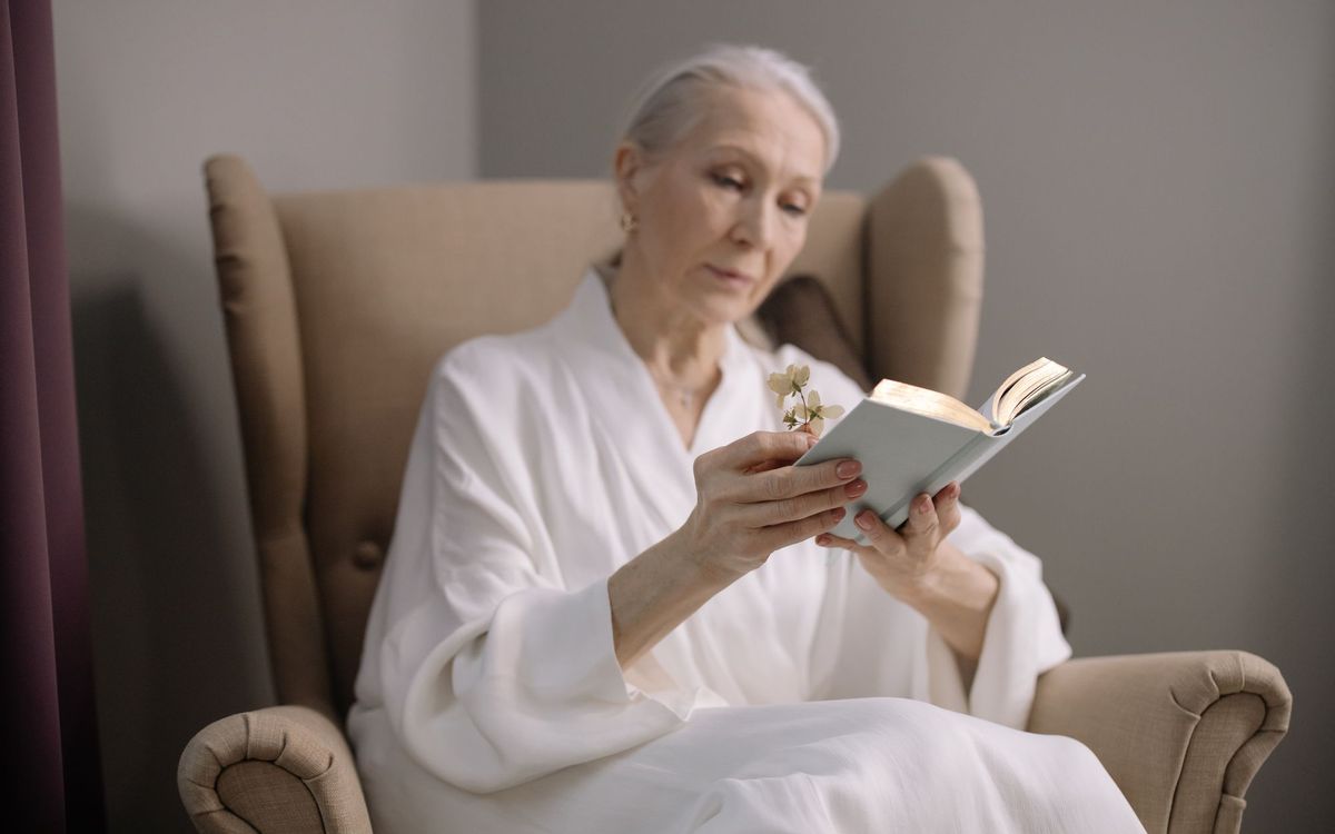 Older woman in a white robe reading a book in a tan wingback armchair
