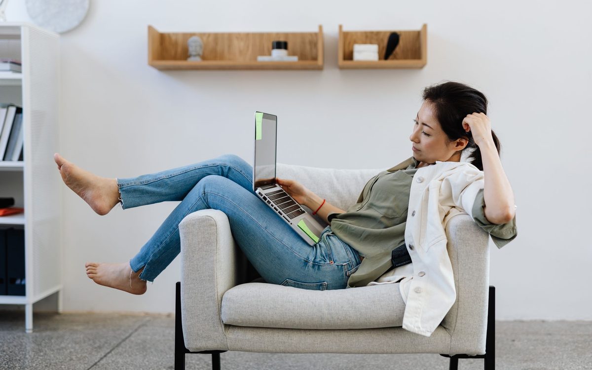 Woman lounging with her legs up in a comfortable armchair beside a white wall while working on a laptop