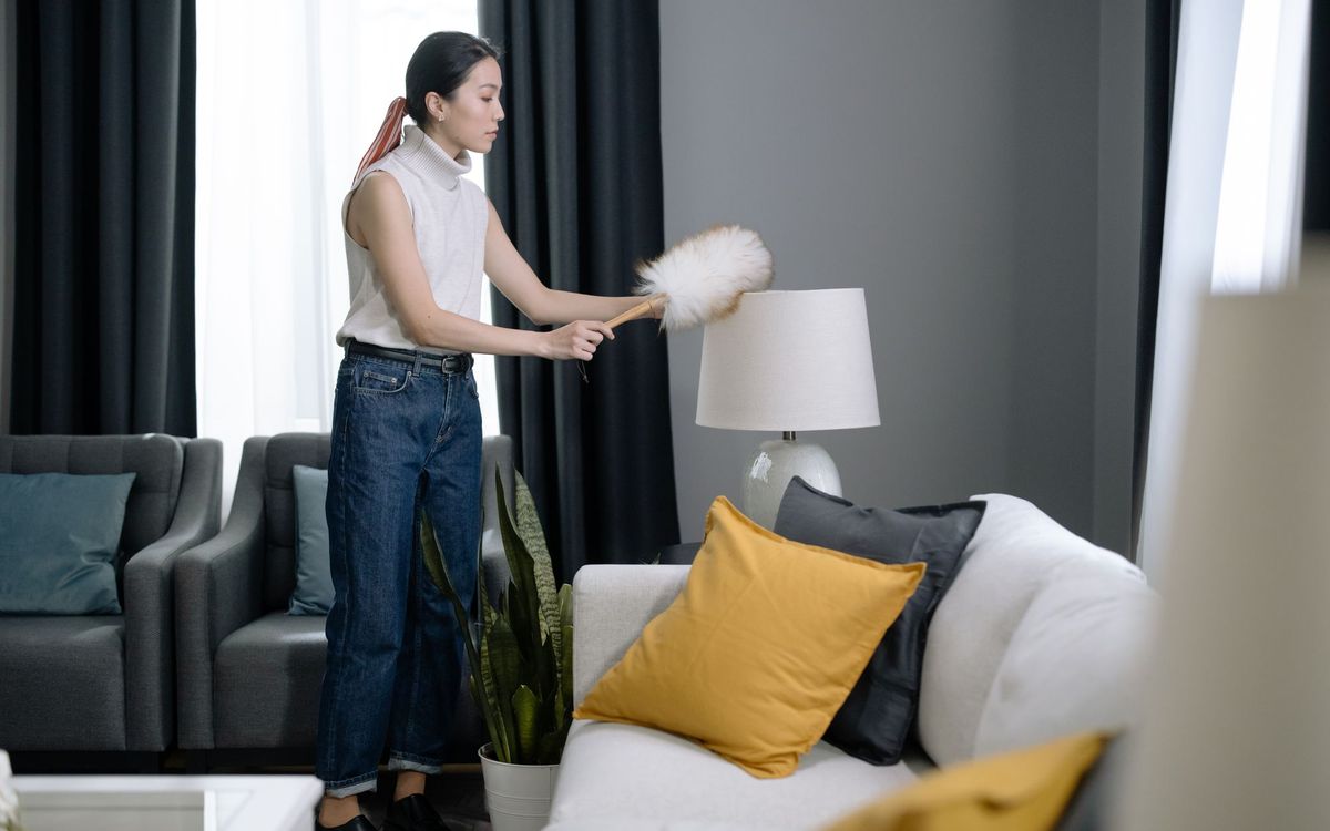 Woman dusting a lamp with a feather duster in a living room with armchairs and a sofa