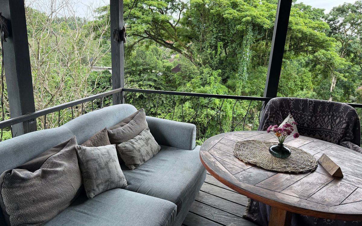 Covered outdoor space with a gray sofa and a weathered round wood table overlooking greenery.