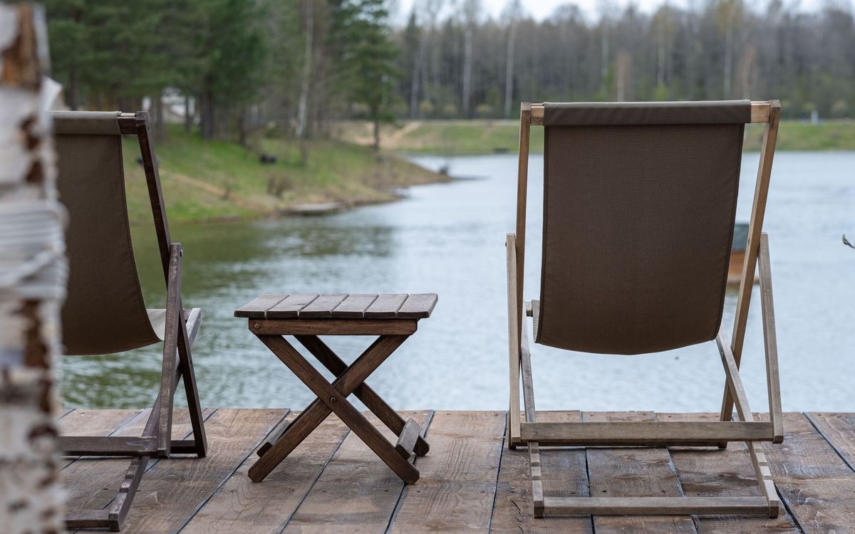 Two wooden folding deck chairs with fabric slings and a small slatted wooden table on a plank deck beside a lake.