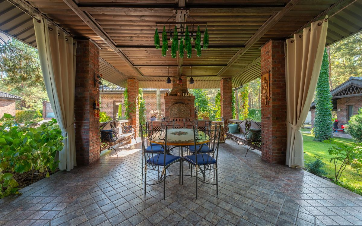 Covered brick patio with a round metal table, metal chairs with blue seat pads, two metal benches and open curtains.