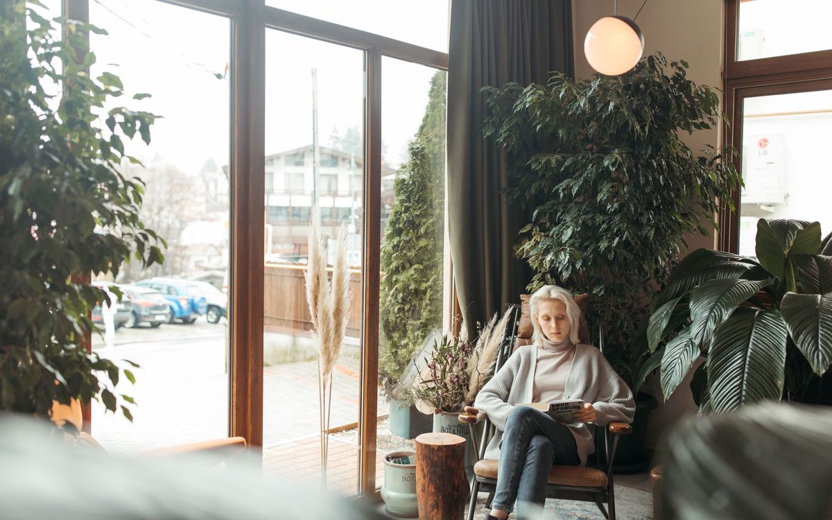 A woman in a cardigan reading a magazine in a wooden armchair beside large windows and plants