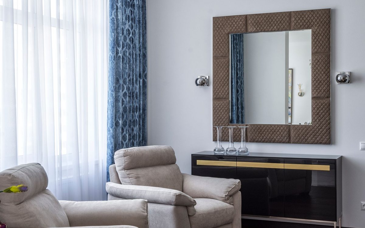 Two soft beige upholstered armchairs beside a white curtain and a mirror in a bright living room